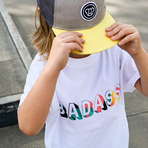 garçon dans un skatepark avec look streetwear casquette snapback pour enfant grise et jaune, t-shirt large oversize unisexe blanc en coton bio, motif Badass coloré