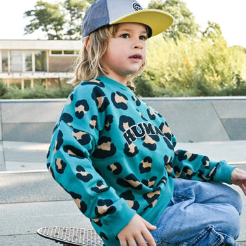 petit garçon dans un skatepark look streetwear cool sur un skate avec un sweat oversize unisexe en coton bio motif léopard bleu vert et une casquette snapback pour enfant visière plate grise et jaune