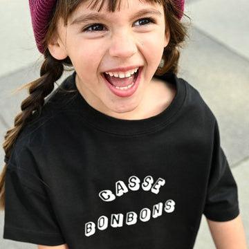 petite fille souriante portant un t-shirt unisexe streetwear large noir, en coton bio, avec motif Casse Bonbons et un bonnet pour enfant en laine couleur prune