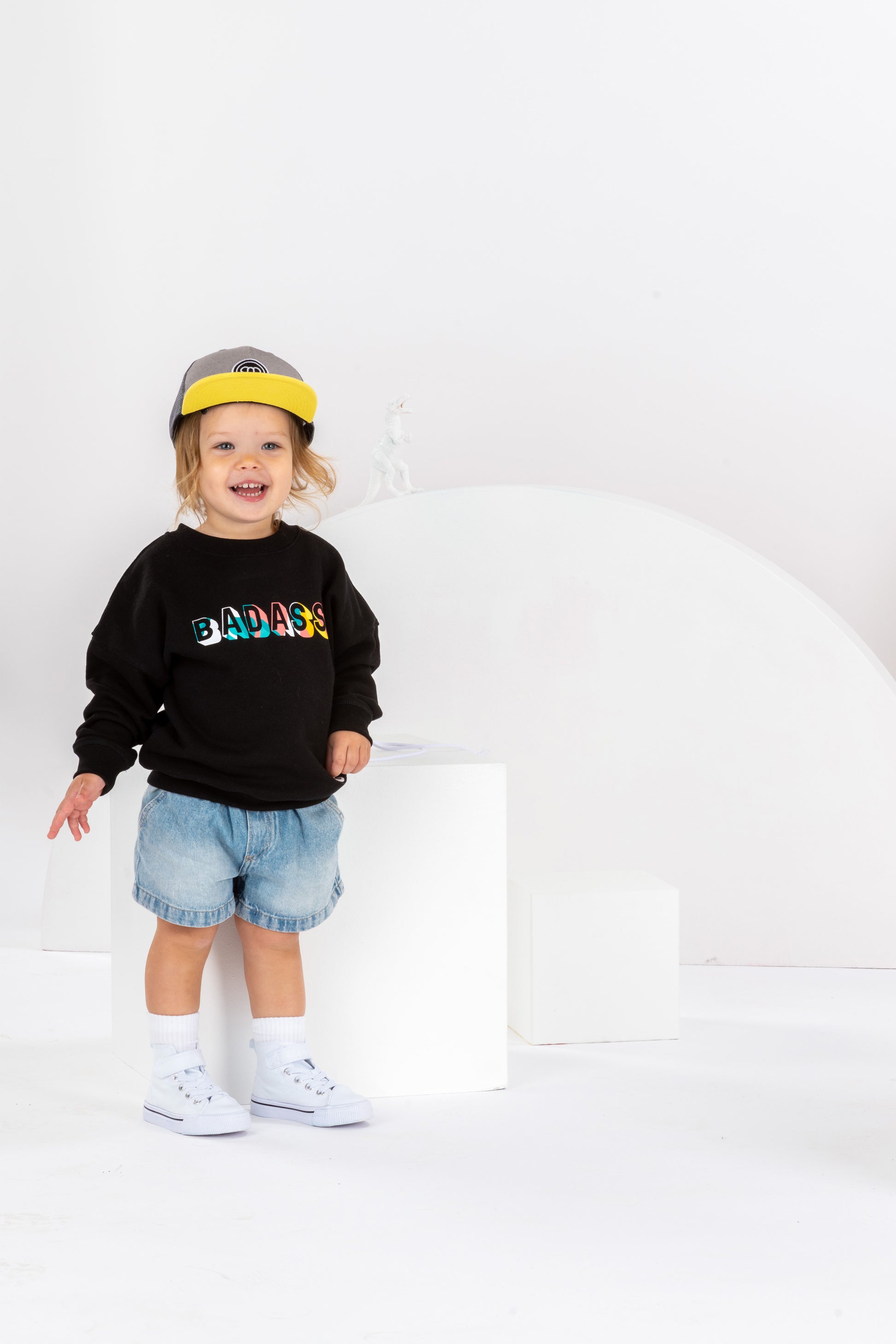 petite fille souriante de 2 ans dans un décor monochrome blanc, en tenue streetwear cool, sweatshirt noir pour enfant imprimé Badass en coton bio, coupe unisexe oversize, casquette réglable pour enfant grise et jaune, visière plate, short en jean, baskets montantes blanches