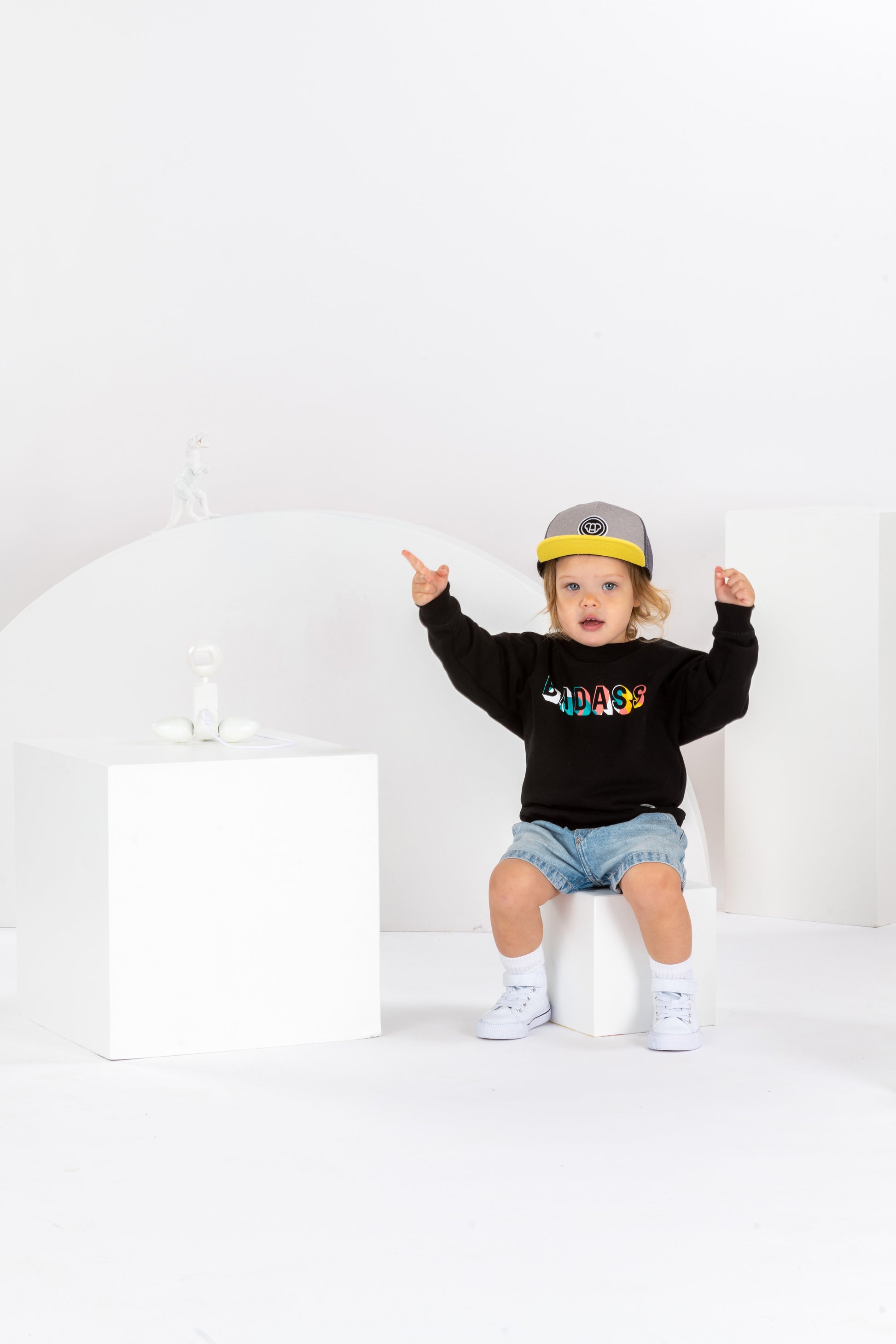 petite fille de 2 ans, assise dans un décor monochrome blanc, en tenue streetwear cool, sweatshirt noir pour enfant imprimé Badass en coton bio, coupe unisexe oversize, casquette réglable pour enfant grise et jaune, visière plate, short en jean, baskets montantes blanches