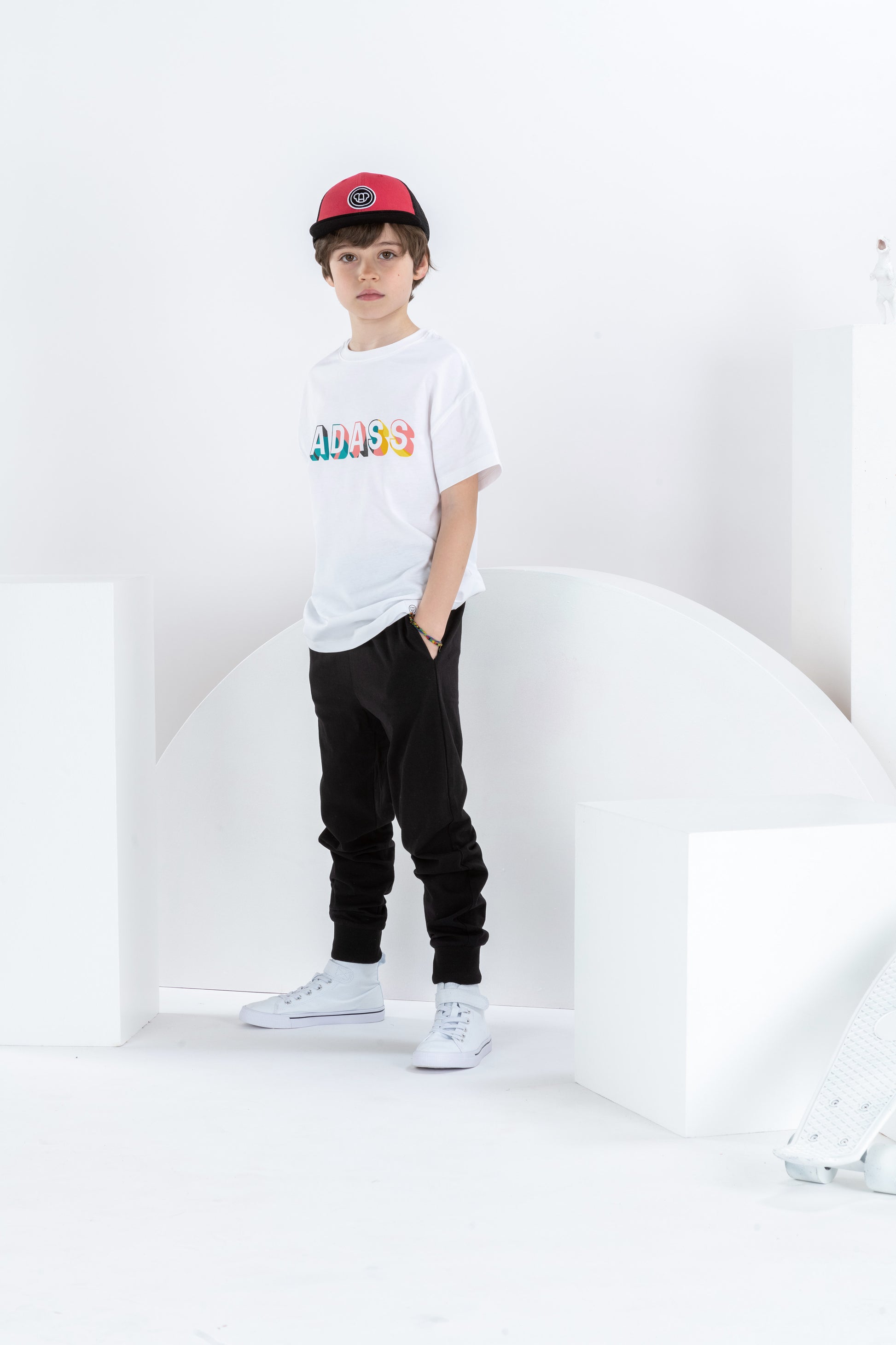 garçon de 8 ans dans un décor monochrome blanc avec un skateboard, en tenue streetwear cool avec t-shirt blanc pour enfant, en coton bio, coupe oversize unisexe, motif cool imprimé Badass coloré, casquette réglable rouge et noire pour enfant, pantalon jogging noir et baskets blanches montantes