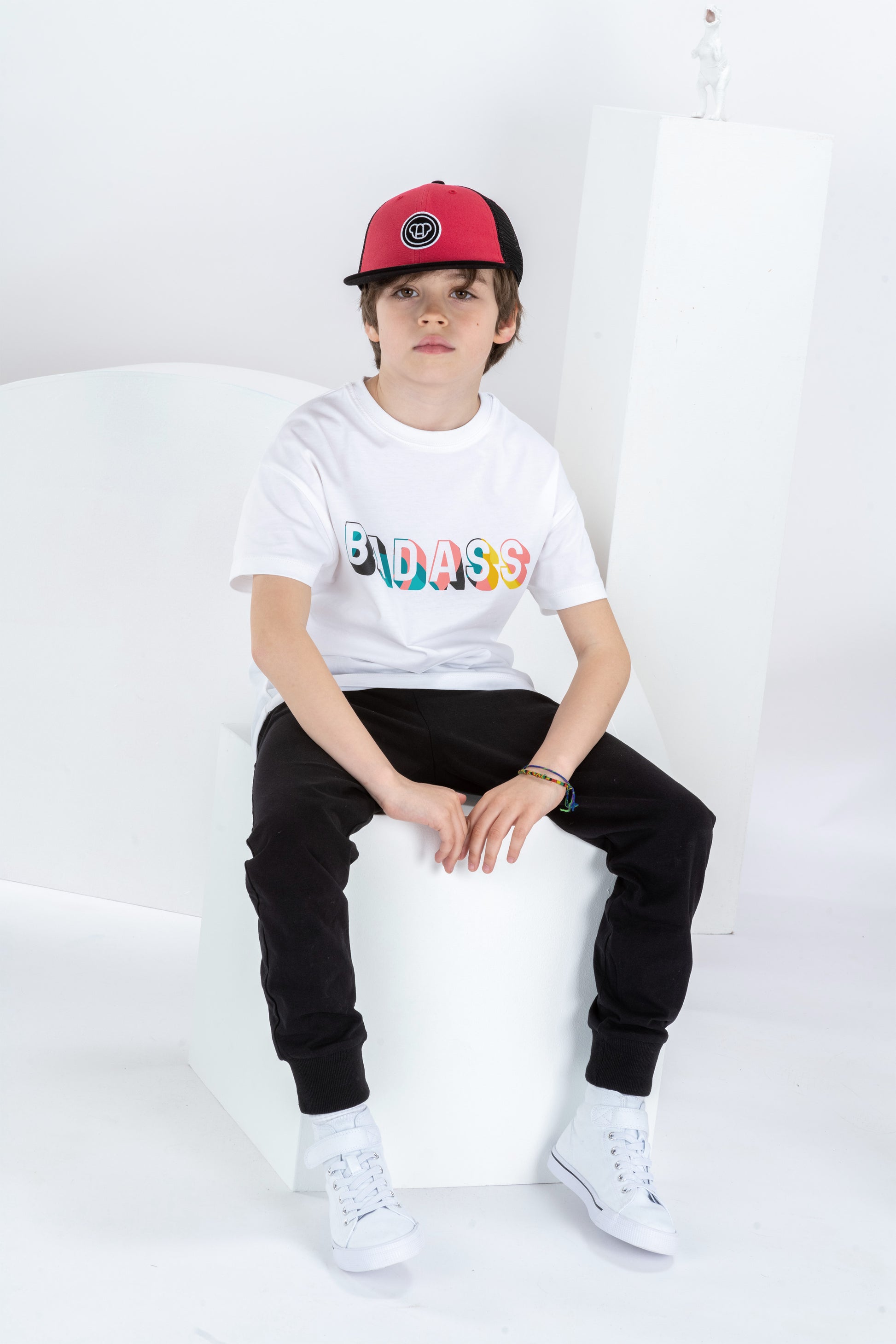 garçon de 8 ans assis dans un décor monochrome blanc, tenue streetwear cool avec t-shirt blanc pour enfant, en coton bio, coupe oversize unisexe, motif cool imprimé Badass coloré, casquette réglable rouge et noire pour enfant, pantalon jogging noir et baskets blanches montantes