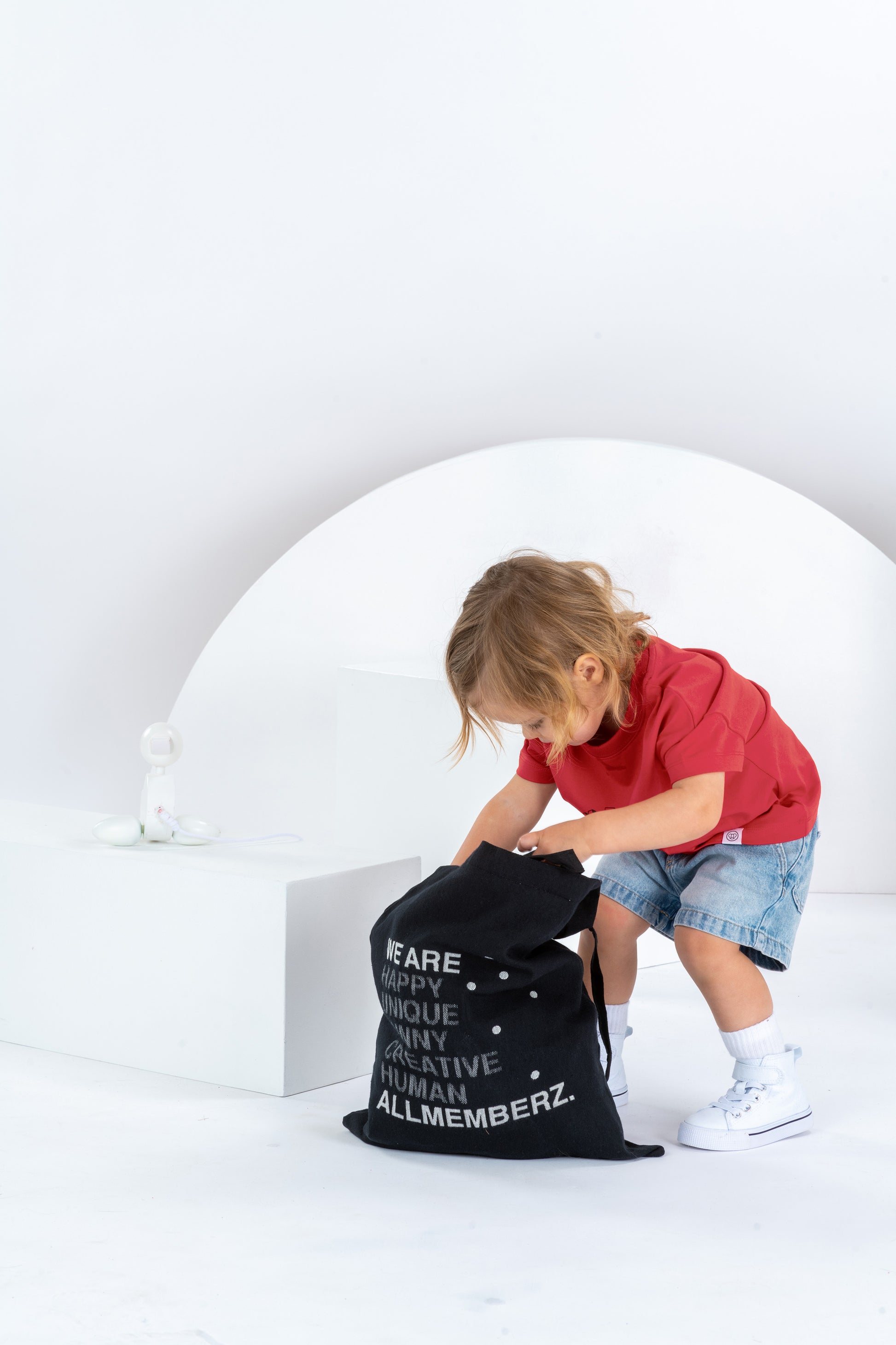 Petite fille de 2 ans cherchant dans un sac en toile tote bag noir Allmemberz avec message graphique, dans un décor monochrome blanc